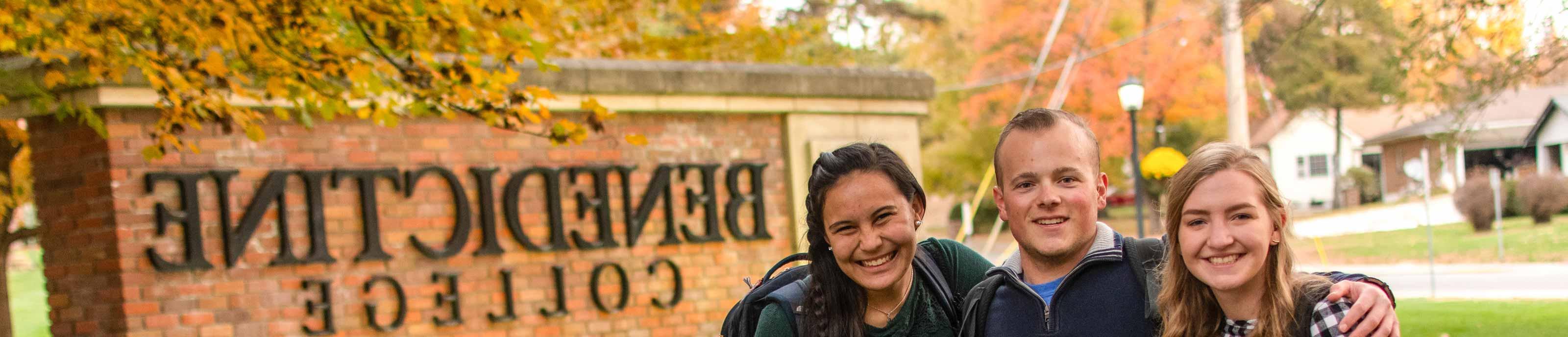 Three students pose for a photo in front of the Benedictine College entrance sign